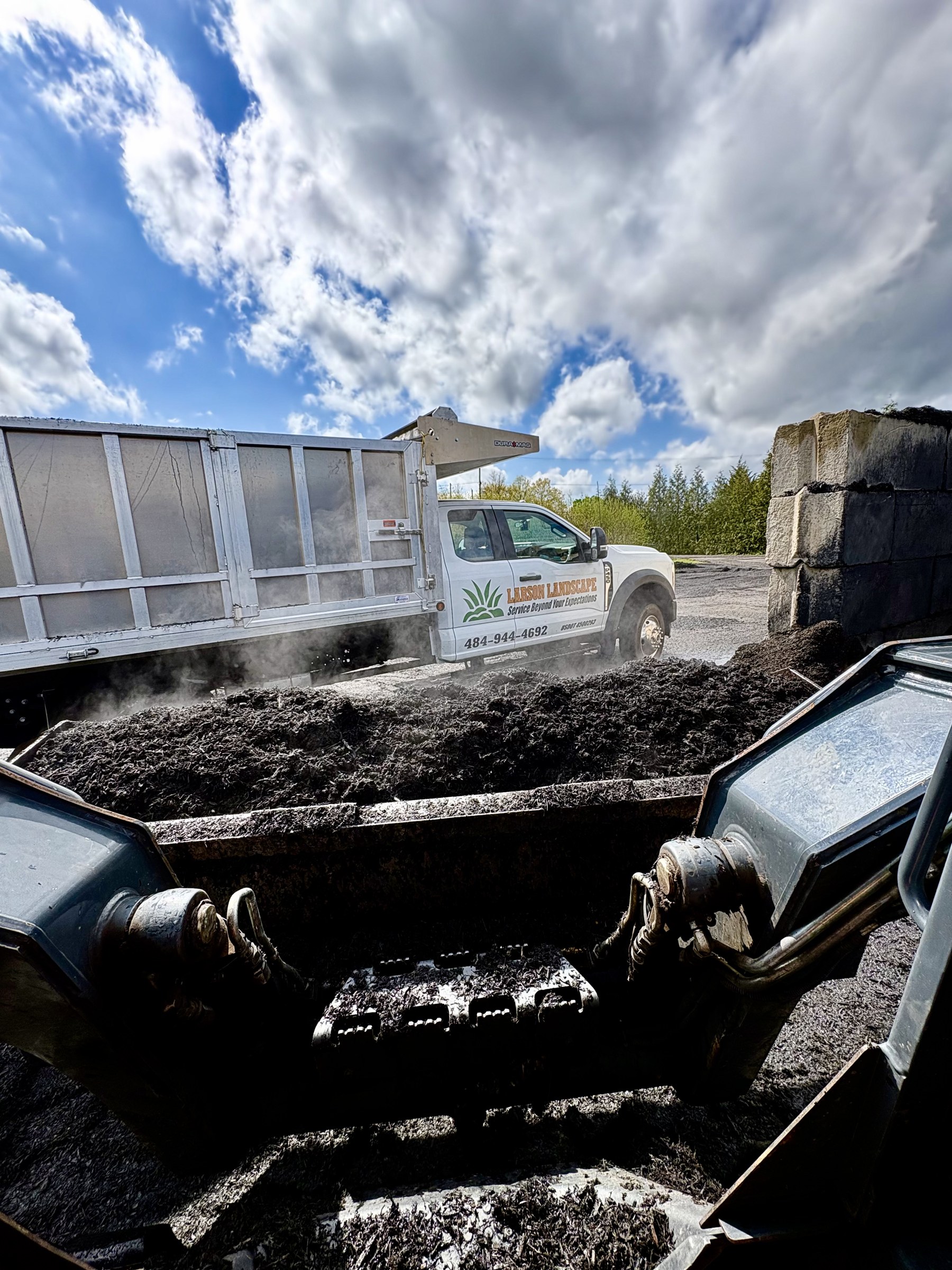 Larson Landscape branded dump truck loading fresh, steaming mulch with a skid steer bucket